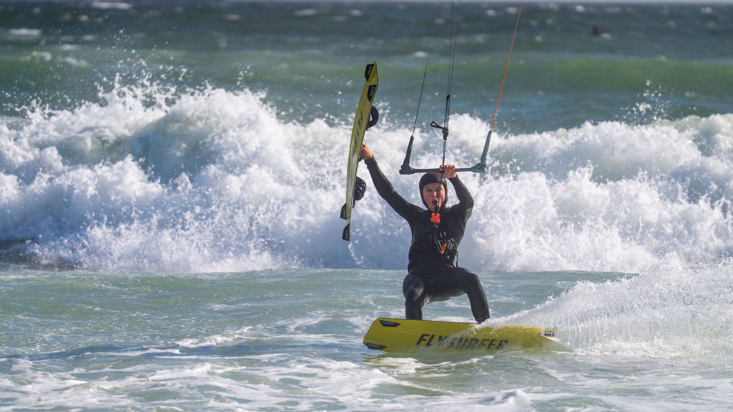 Kajtar na deski Flysurfer SHOT med vožnjo po valovih – akcijski prizor z morja v realnih pogojih za športno navdušene popotnike.