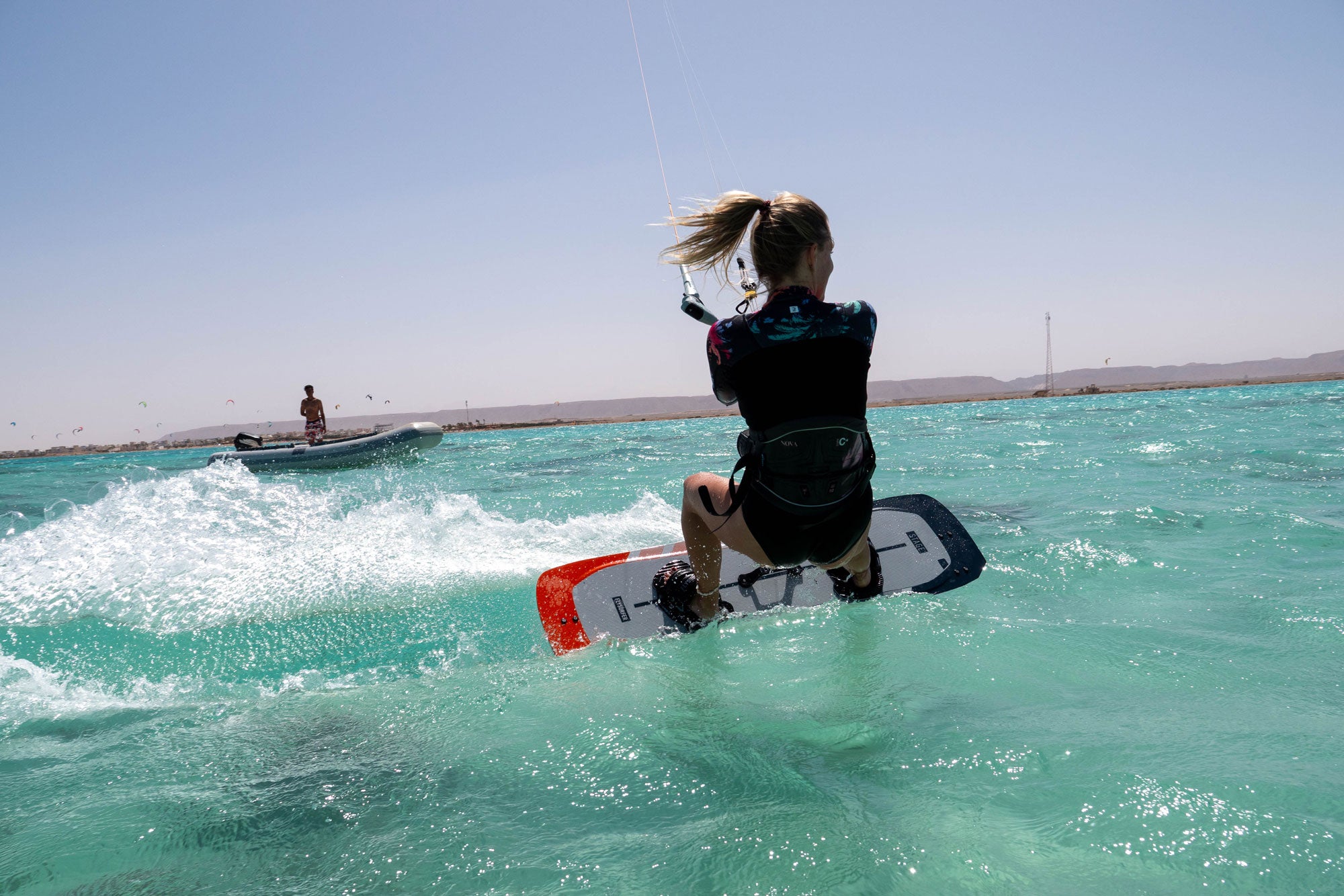 Kajtarka na deski Flysurfer med vožnjo po turkiznem morju ob vetrovnem dnevu v Egiptu; v ozadju je gumenjak z inštruktorjem in puščavska obala.