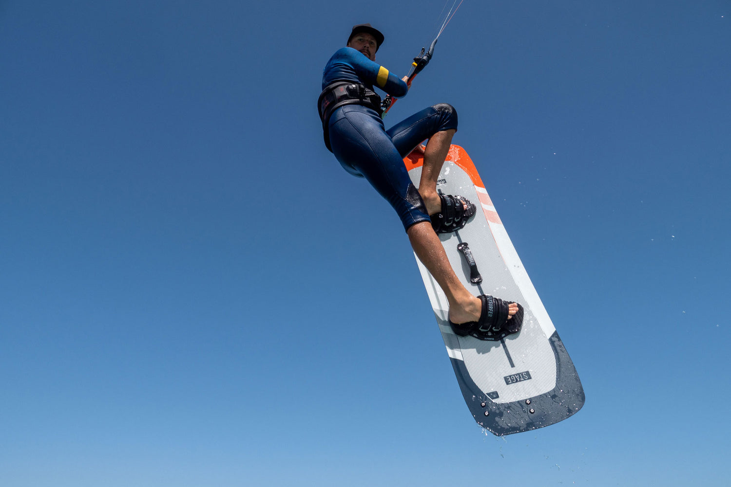 Kitesurfer v zraku z desko Flysurfer Stage med trikom na jasnem modrem nebu.