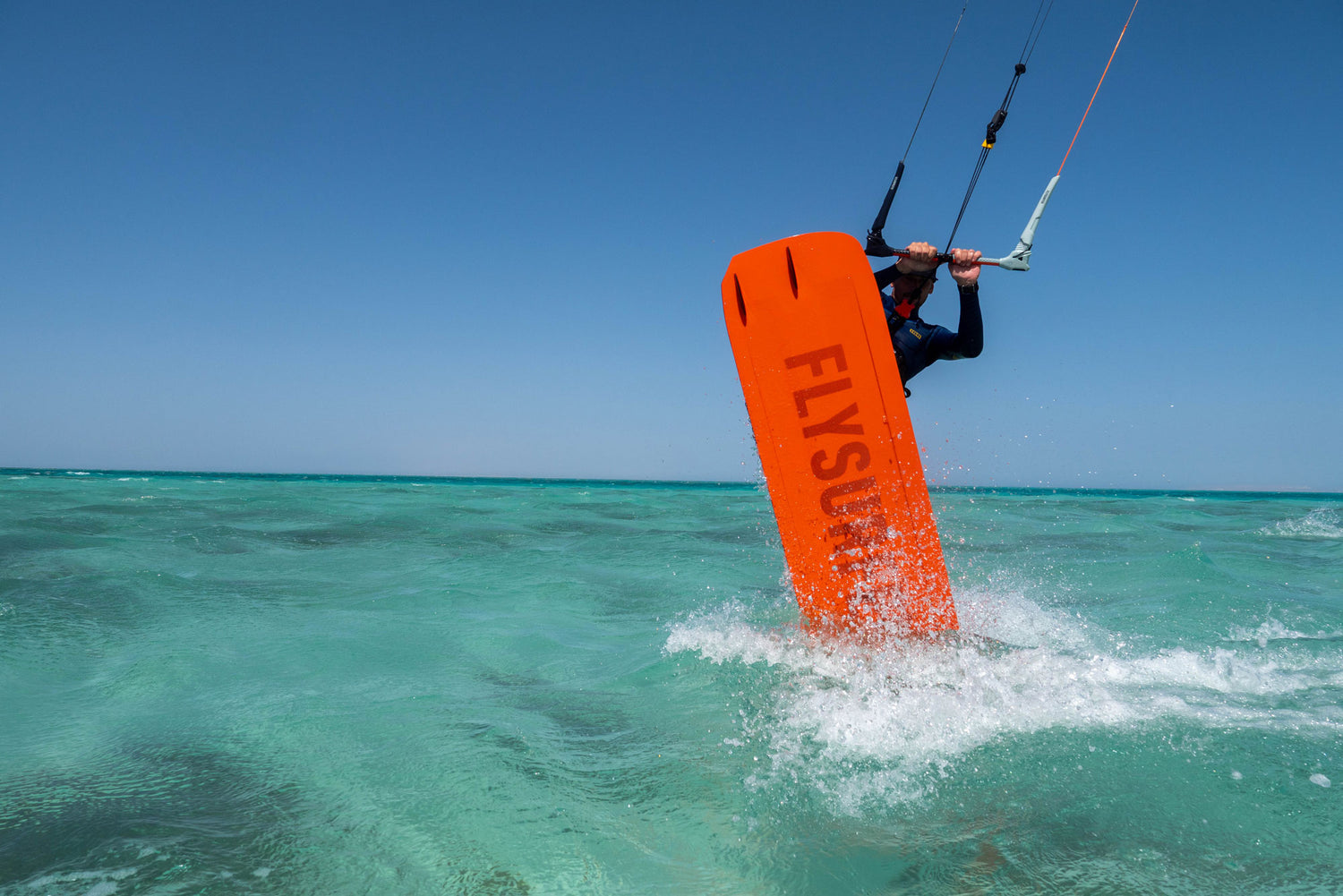 Kajtarska akrobacija z oranžno desko Flysurfer na kristalno čistem morju pod modrim nebom.