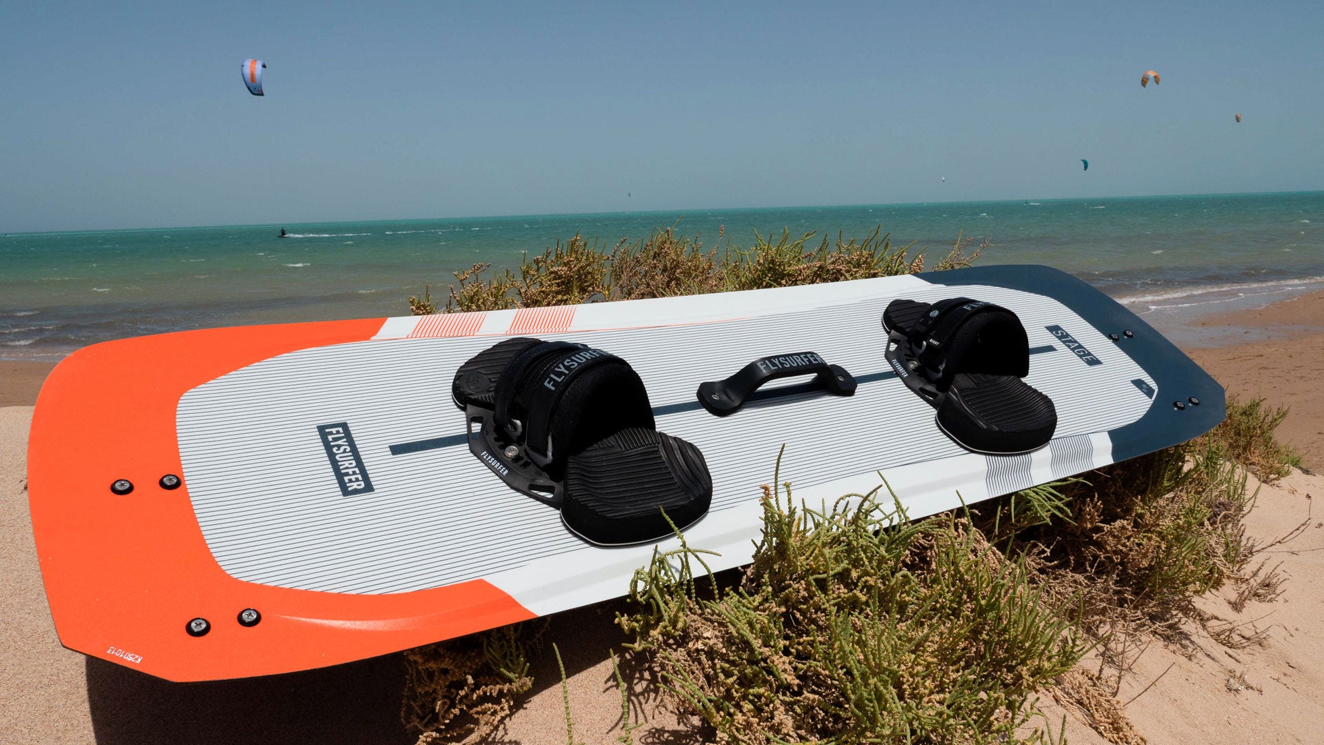 Flysurfer kiteboard z naramnimi paščki, pripravljen na vožnjo, fotografiran na peščeni plaži ob morju z vetrom v ozadju – idealen za kajtanje na vodi.