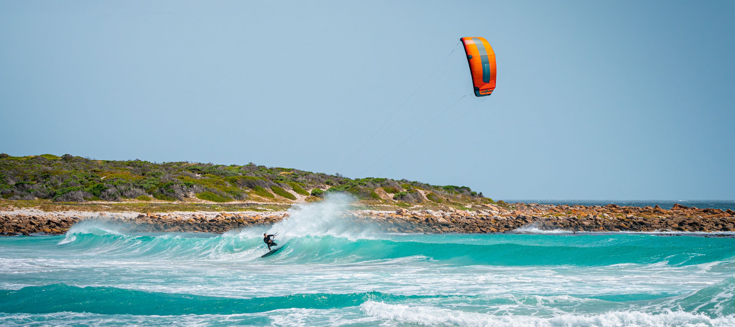 Kitesurfer deska po turkiznih valovih ob sončnem dnevu na osamljeni plaži z zaraščeno obalo v ozadju. Športna avantura na morju.
